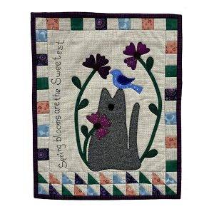 wall hanging featuring a cat with flowers with the words spring blloms are the sweetest embroidered on it