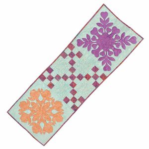 Long cotton table runner with Hawaiian appliqué designs of palm fronds, pineapples, and hibiscus in pastel colors on a light background. Measures 18x54 inches.