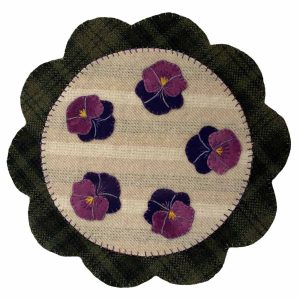 Circular wool mat with five hand-embroidered purple and green pansy flowers on a light tan textured background. Measures 11.5 x 11.5 inches.